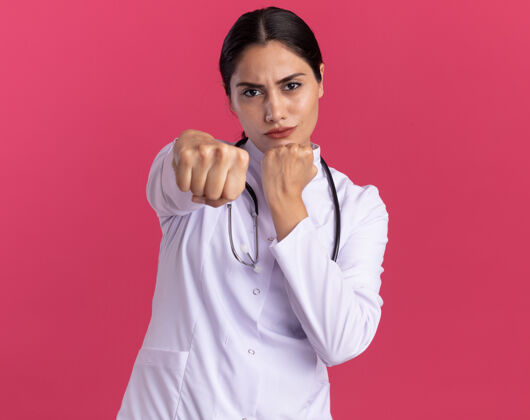 脸年轻的女医生 穿着医用外套 手持听诊器 正脸严肃地站在粉红色的墙上 面前握紧拳头外套医生秀