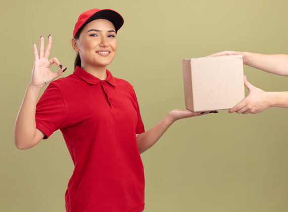 盒子身着红色制服 头戴鸭舌帽的年轻女送货员微笑着向站在绿色墙壁上的顾客赠送纸板箱 上面贴着“ok”标志送货女人朋友