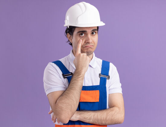 站立年轻的建筑工人穿着建筑制服 戴着安全帽 站在紫色的墙上 用食指指着前面的眼睛 感到困惑目录建筑眼睛