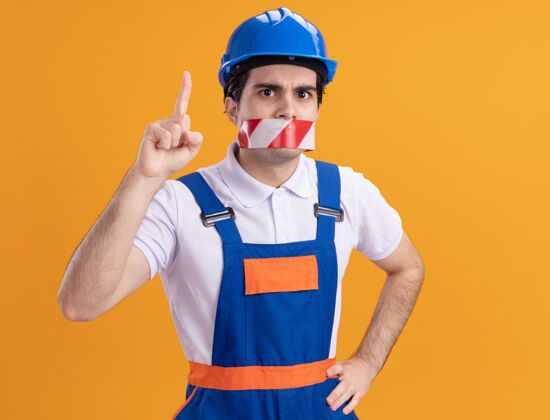 年轻人年轻的建筑工人穿着建筑制服 戴着安全帽 嘴上缠着胶带 正焦急地看着前面 食指朝上 站在橙色的墙上担心指向嘴