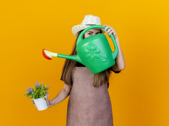 花看着镜头美丽的园丁女孩穿着制服 戴着园艺帽 手里拿着花盆里的花 脸上沾满了水 可以隔离在橙色的背景上相机浇水花园
