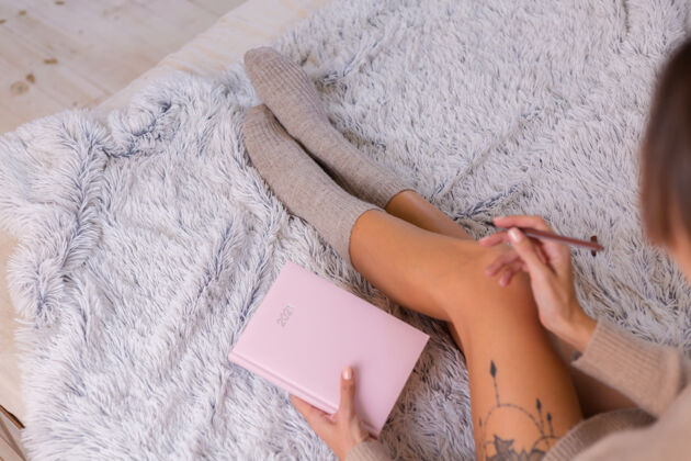 日记穿着羊毛袜子和毛衣的女人 粉色笔记本上写着2021 臀部有大纹身女人坐在卧室的家里的床上计划者腿温暖