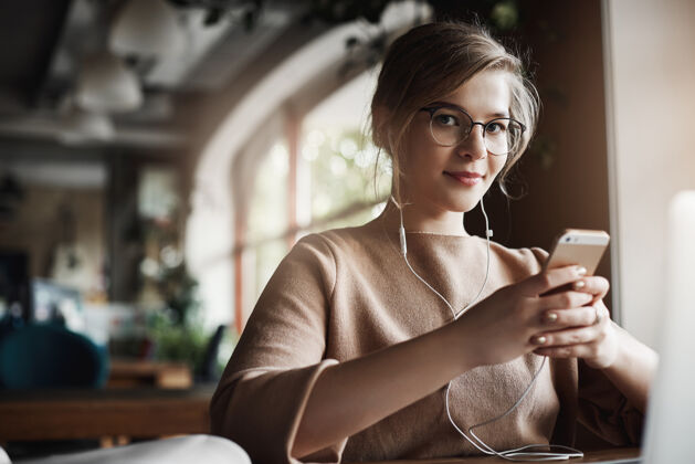 人室内拍摄的漂亮时尚快乐的白人女性 金发戴眼镜 手持智能手机 戴着耳塞 一边看视频 分心地看着和微笑设备人笔记本电脑