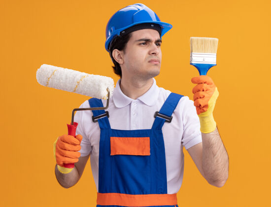 混乱年轻的建筑工人穿着建筑制服 戴着安全帽 戴着橡胶手套 手里拿着油漆辊和刷子 站在橙色的墙上 看上去很困惑橡皮手握男人