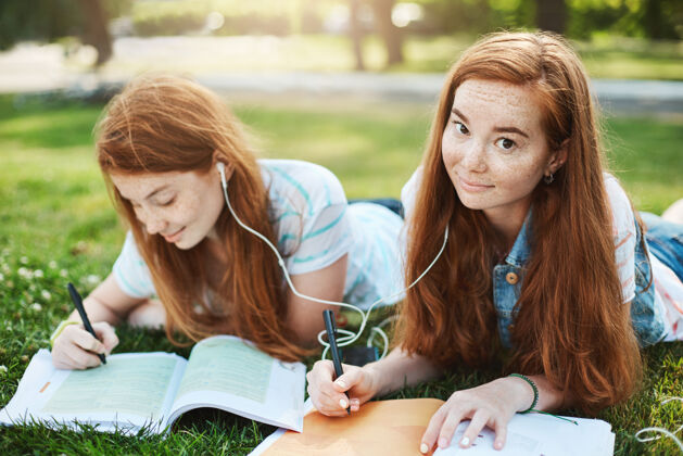 红发姜黄色的头发 扬起眉毛和可爱的微笑 躺在草地上 在城市公园与妹妹 分享耳机一起听音乐和做作业生活方式和人的概念朋友耳塞草