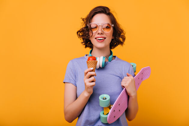 多彩微笑的白人女孩拿着长板吃甜点的肖像黑发迷人的女人拿着滑板和冰淇淋室内快乐欢呼
