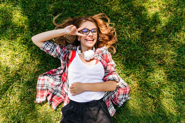 可爱戴着白色耳机的快乐女孩躺在草地上 面带微笑户外俯瞰拍摄的温文尔雅的女人在草坪上发冷年轻放松成人