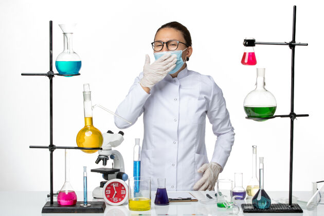人前视图：身穿医疗服的女化学家 戴着面具 打着哈欠 背景是白色的病毒化学实验室的covid-splash研究医学前面