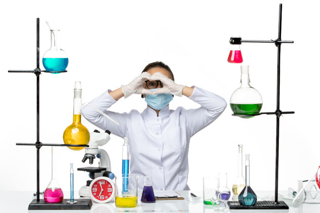 化学前视图穿着医疗服的女化学家带着面罩坐在桌子前 溶液背景为浅白色病毒化学实验室的covidsplash坐面具专业