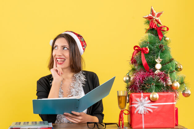 西装穿着西装 戴着圣诞老人帽和新年装饰品的梦幻商务女士正在检查文件 坐在一张桌子旁 桌子上放着圣诞树圣诞老人年文件