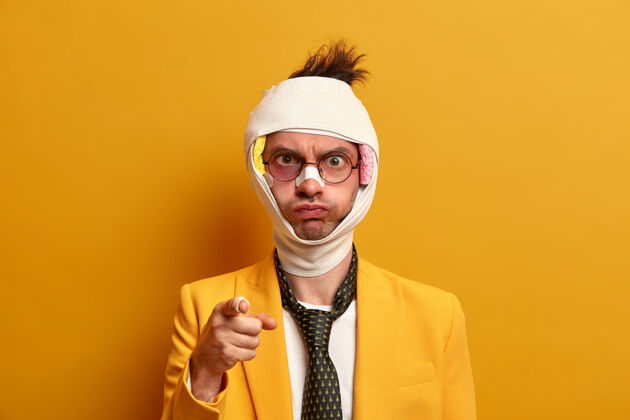 姿势愤怒的受伤住院男子指着并责怪有人对他的事故 脑震荡 包扎头部 穿着正式 隔离在黄色墙壁上 需要治疗创伤骨折事故