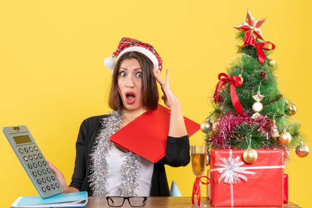 计算器穿着西装 戴着圣诞老人帽和新年装饰品 手里拿着计算器 坐在一张桌子旁 桌子上放着圣诞树桌子成人快乐