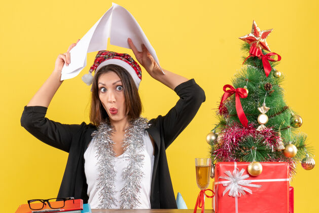 圣诞老人情绪化的商务女士穿着西装 戴着圣诞老人的帽子 戴着新年装饰品 头上举着文件 坐在一张桌子旁 桌子上放着圣诞树头西装文件