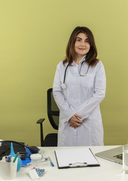 立场高兴的中年女医生穿着医用长袍和听诊器站在办公桌后面 手里拿着医疗工具剪贴板和笔记本电脑 双手分开中年医疗桌子
