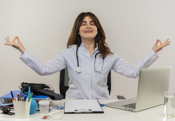 听诊器一位身穿医用长袍 听诊器的中年女医生坐在办公桌旁 手里拿着医疗工具 剪贴板和笔记本电脑 微笑着 闭眼沉思眼睛剪贴板成人