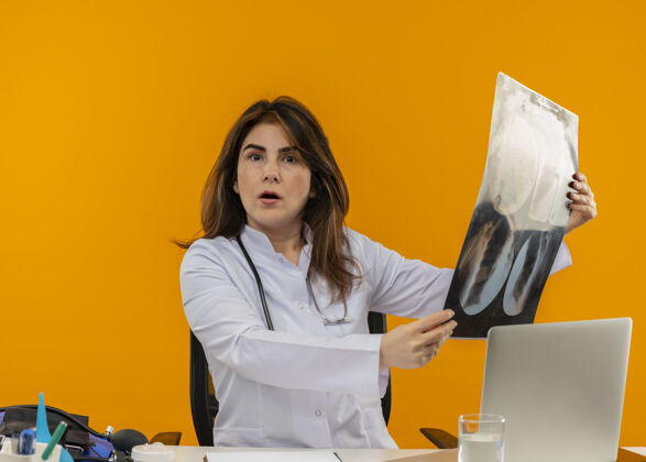 拿着令人印象深刻的中年女医生穿着医用长袍和听诊器坐在办公桌旁用医疗工具剪贴板和笔记本电脑拿着x光片橙色坐着剪贴板
