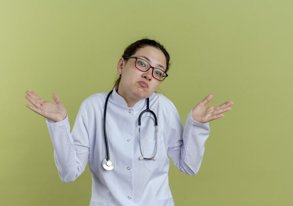 绿色困惑的年轻女医生穿着医用长袍和听诊器 戴着眼镜 双手分开医学传播眼镜