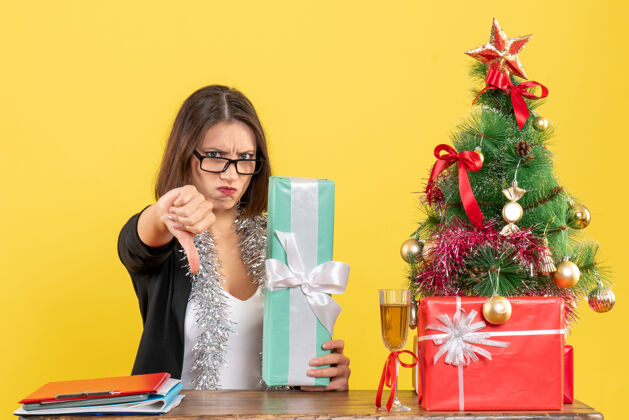 展示一位穿着西装戴着眼镜的漂亮女士在办公室里摆着一张桌子 桌上放着一棵圣诞树成人礼物手势
