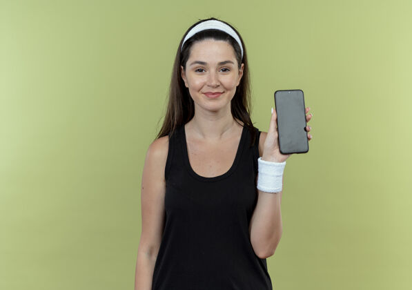 站立戴着头巾的年轻健身女士展示着智能手机 自信地微笑着看着站在灯光背景下的相机移动自信头带