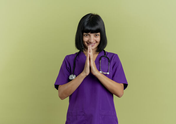 制服带着微笑的年轻黑发女医生穿着制服 手持听诊器 在橄榄绿的背景上 双手分开 留有复印空间年轻持有一起