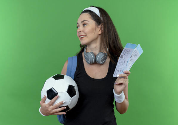 健身戴着头戴耳机 背着背包 拿着足球和机票 微笑着站在绿色背景下的年轻健身女士背包机票空气