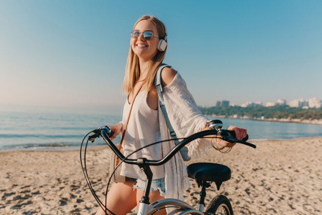 放松年轻迷人的金发微笑的女人 戴着耳机骑着自行车走在沙滩上听音乐女性阳光旅行者