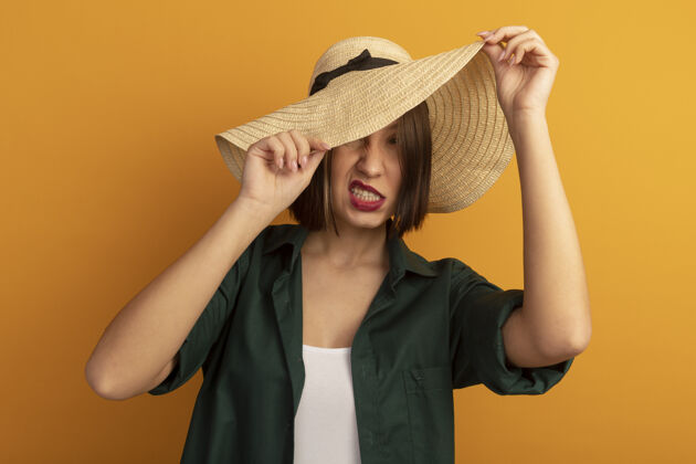 掩护恼怒的美女用沙滩帽遮住脸 隔离在橙色的墙上烦恼表情脸