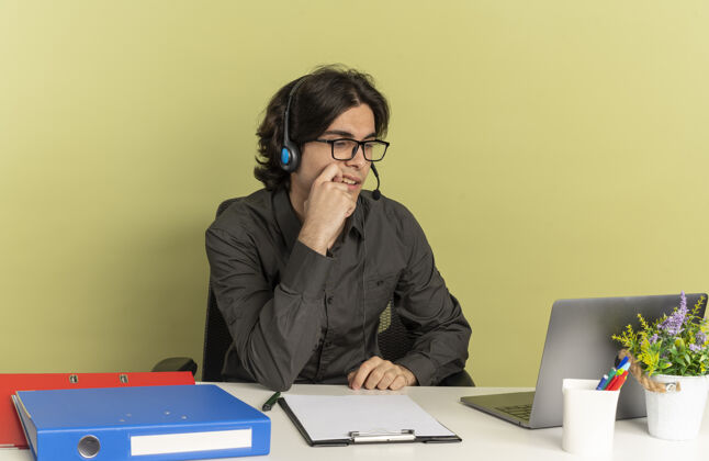 办公桌年轻自信的上班族戴着耳机 戴着眼镜 坐在办公桌旁 手里拿着办公工具 看着笔记本电脑 手放在下巴上 隔离在绿色背景上 留着复印空间光学坐着使用