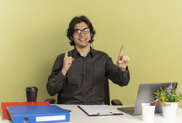 坐着戴着耳机 戴着眼镜 面带微笑的年轻上班族坐在办公桌旁 手里拿着办公工具 手里拿着笔记本电脑 在绿色背景上点着独立的复印空间工具工人使用