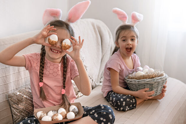 节日有兔子耳朵的小姐妹们 家里的沙发上放着复活节彩蛋鸡蛋准备童年