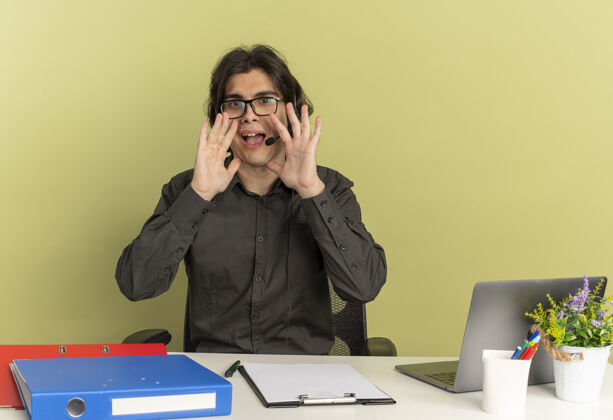 有人戴着耳机 戴着眼镜 坐在办公桌旁 拿着办公工具 用笔记本电脑的年轻快乐的上班族 假装在绿色背景下给一个有复印空间的人打电话工人办公桌笔记本电脑