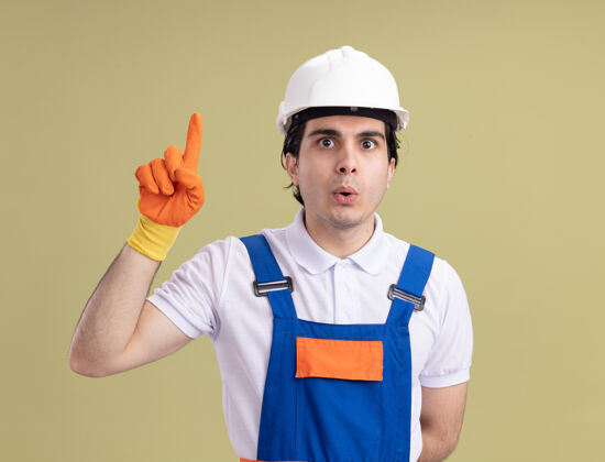 建筑年轻的建筑工人穿着建筑制服 戴着安全帽 戴着橡胶手套 惊讶地看着前面 伸出食指 站在绿色的墙上 有了新的想法手指橡胶手套