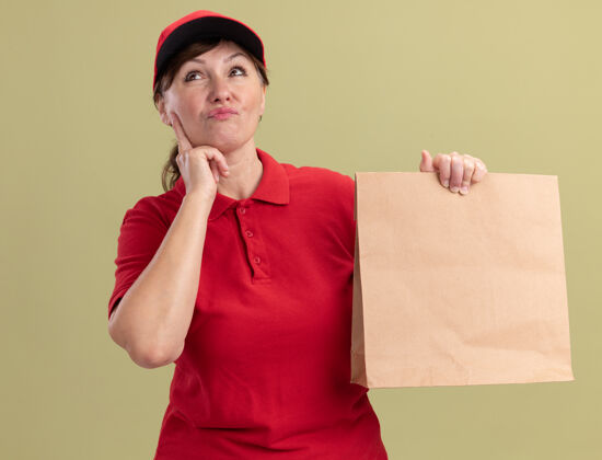 沉思身穿红色制服 戴着礼帽的中年送货妇女站在绿色的墙壁上 脸上带着沉思的表情抬起头来思考包装女人思考