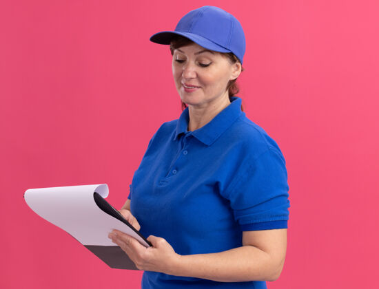 帽子身穿蓝色制服 戴着帽子的中年送货妇女站在粉色的墙上 拿着一本空白的剪贴板 面带微笑地看着它页年龄空白