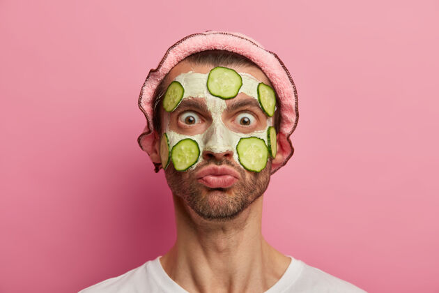 阳刚面部护理概念震惊的情绪化男人盯着他的倒影 用黄瓜片涂上白土面膜面部护理肤色健康
