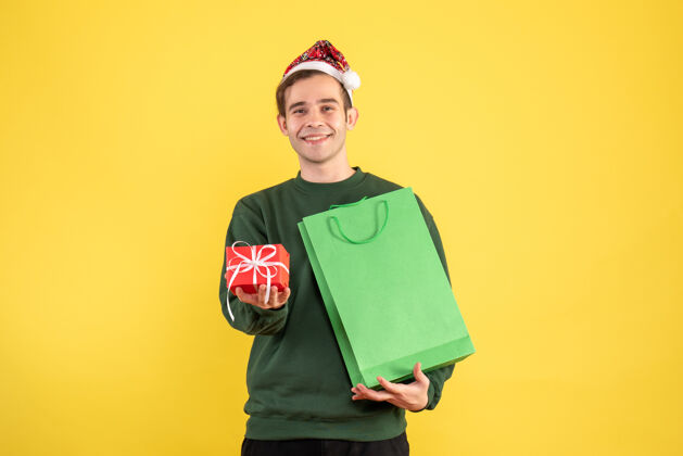 男人正面图戴着圣诞帽的年轻人拿着绿色的购物袋和礼物站在黄色的背景复制空间持有圣诞老人绿色