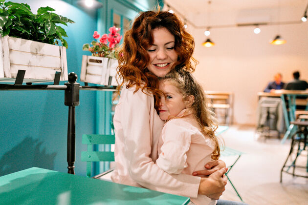 拥抱快乐的年轻女子坐在咖啡馆和她迷人的小女孩拥抱她孩子一起女儿