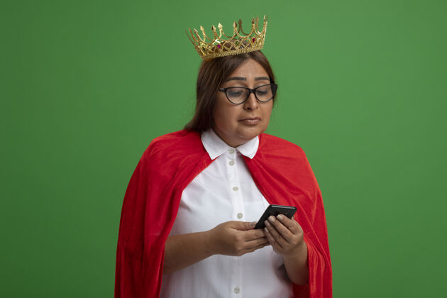 中年高兴的中年女超人戴着皇冠 戴着眼镜 看着绿色背景上孤立的手机手持女皇冠
