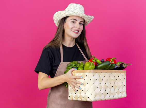 篮子微笑美丽的园丁女孩穿着制服 戴着园艺帽 拿着蔬菜篮子 在粉红色的背景上隔离开来花园微笑制服