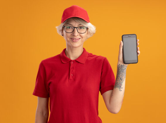 手机身着红色制服 头戴眼镜 站在橙色墙壁上的快乐的年轻送货员送货女士展示