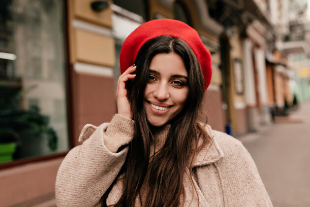 户外老城背景下 一个戴着优雅帽子的无忧无虑的女孩在镜头前摆姿势一个穿着米黄色外套的幸福长发女人在街上笑的户外写真积极时尚淑女