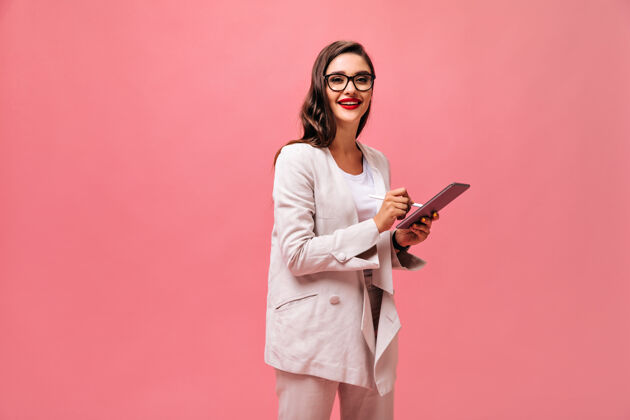 女士穿着时髦夹克 戴着眼镜 红唇的漂亮女人 在孤立的粉色背景下 与电脑平板合影时尚肖像卷发