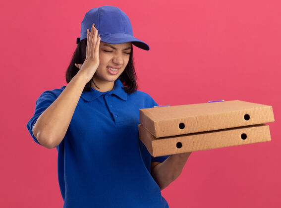 披萨穿着蓝色制服 戴着帽子 拿着披萨盒的年轻女送货员站在粉红色的墙上 看起来不舒服 摸着头 感觉头痛女孩帽子不适