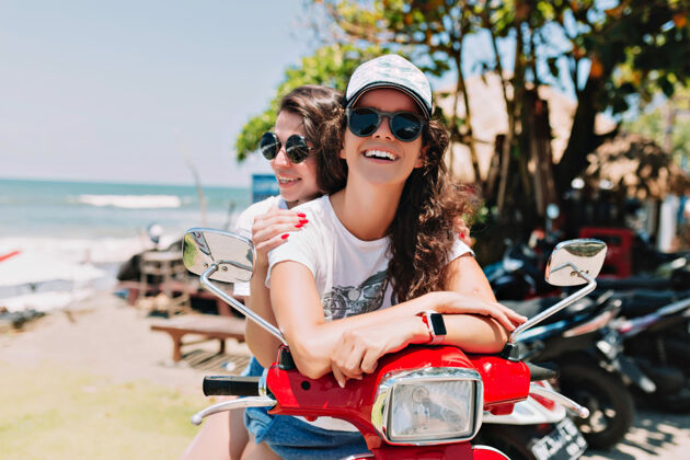 度假村快乐的年轻女性骑着摩托车探索小岛 戴着夏天的帽子 使用平板电脑 在城市背景下在线购买音乐 异国情调的小岛 旅行 暑假享受异国情调女性