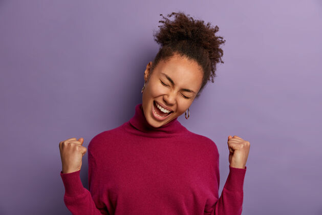 胜利成功兴高采烈的非洲裔美国妇女庆祝惊人的消息 幸运地赢得了很多钱 梦想成真的凯旋 歪着头 穿着休闲的高领毛衣 孤立在紫色的墙上倾斜庆祝年轻