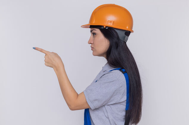 建筑身穿建筑制服 头戴安全帽的年轻建筑妇女站在一边 用食指指着白墙上看起来自信的东西年轻头盔侧面