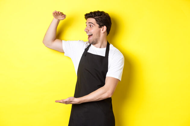 厨师那个男人惊讶地看着一个大东西 站在黄色背景下的黑色围裙里围裙烹饪尺码