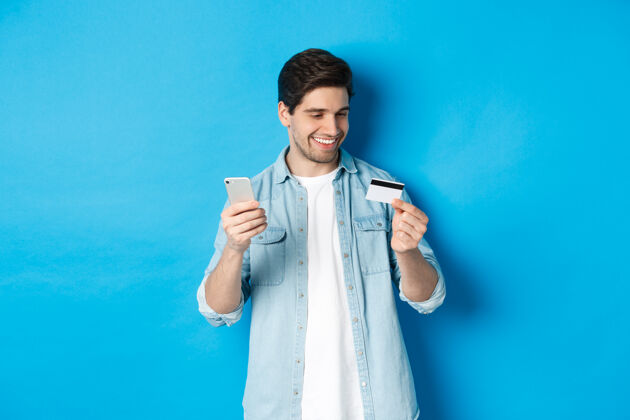 手机英俊快乐的男人在网上购物 拿着信用卡和手机 在网上购物 站在蓝色的背景上信用卡成人工作室