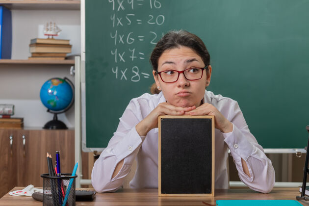 女年轻的女老师戴着眼镜拿着小黑板在教室里讲课 坐在黑板前的课桌上 神情迷茫坐着戴着眼镜