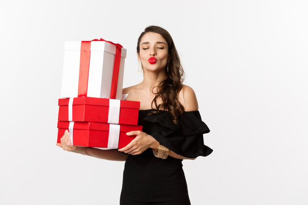亲吻庆祝和圣诞节的概念愚蠢的女人穿着优雅的黑色连衣裙 拿着圣诞节和新年礼物 撅着嘴唇亲吻 快乐地站在白色背景上优雅女性美容